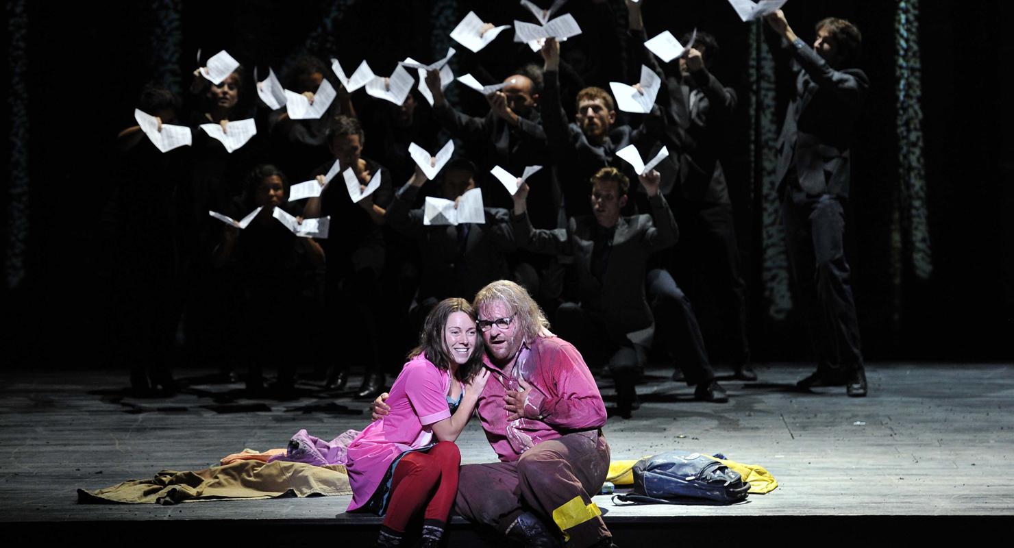 Man and woman in pink shirts sitting while group behind them hold torn pages in ENO's The Magic Flute