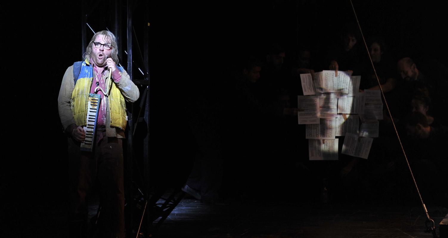 Man playing accordion standing next to papers stuck to a wall in ENO's The Magic Flute