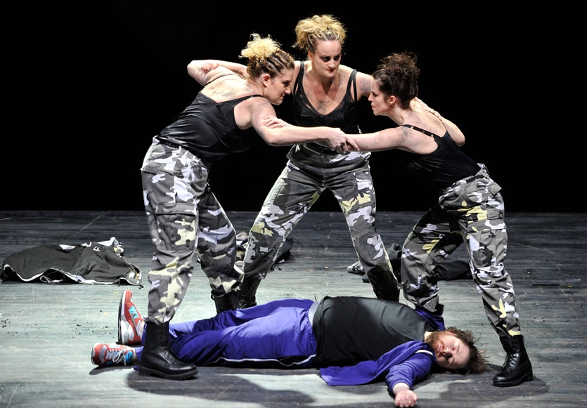 three women in army trousers surrounding man lying on the floor