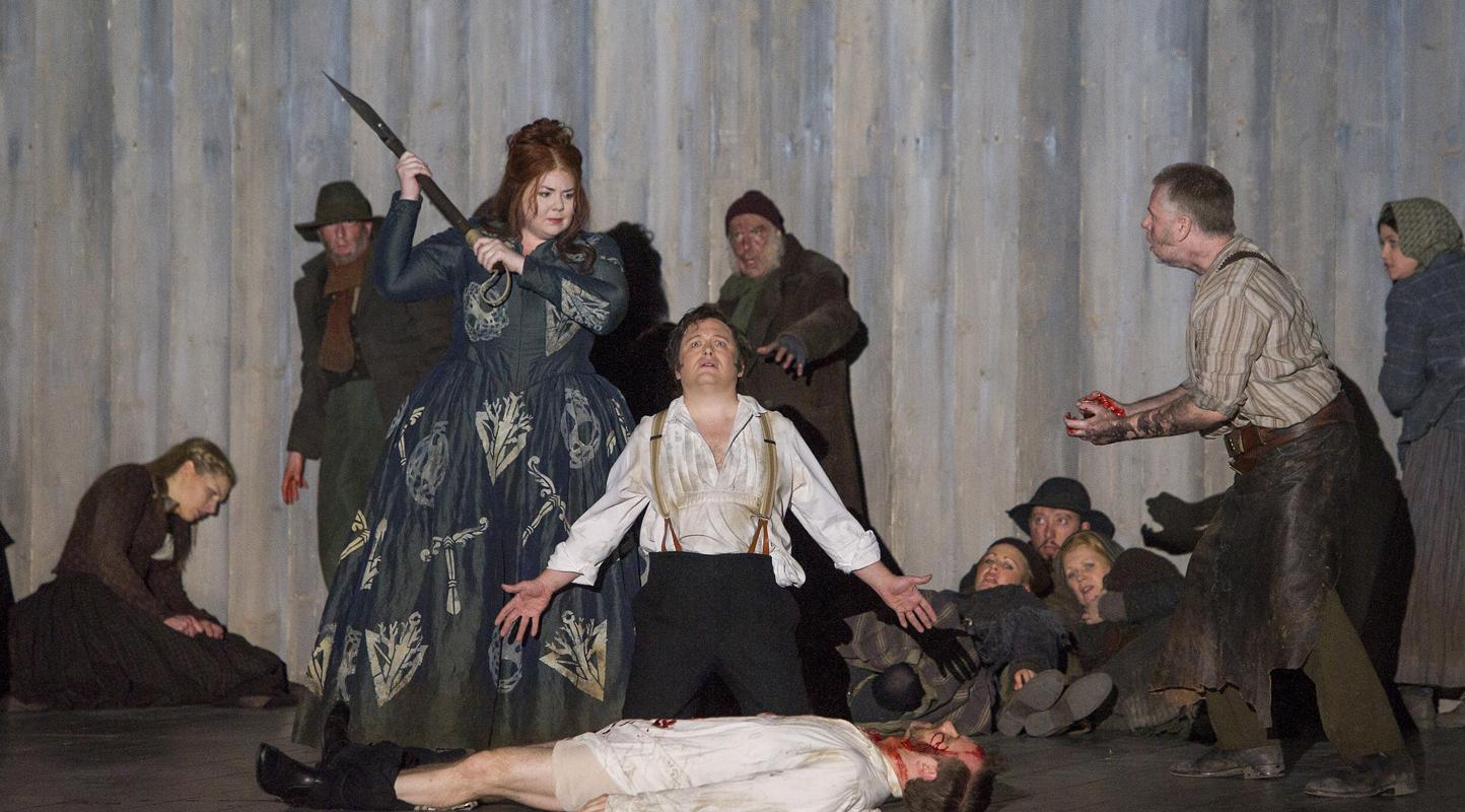 Woman in blue dress holding up a blunt object, while a man lays on the floor dying in ENO's Norma