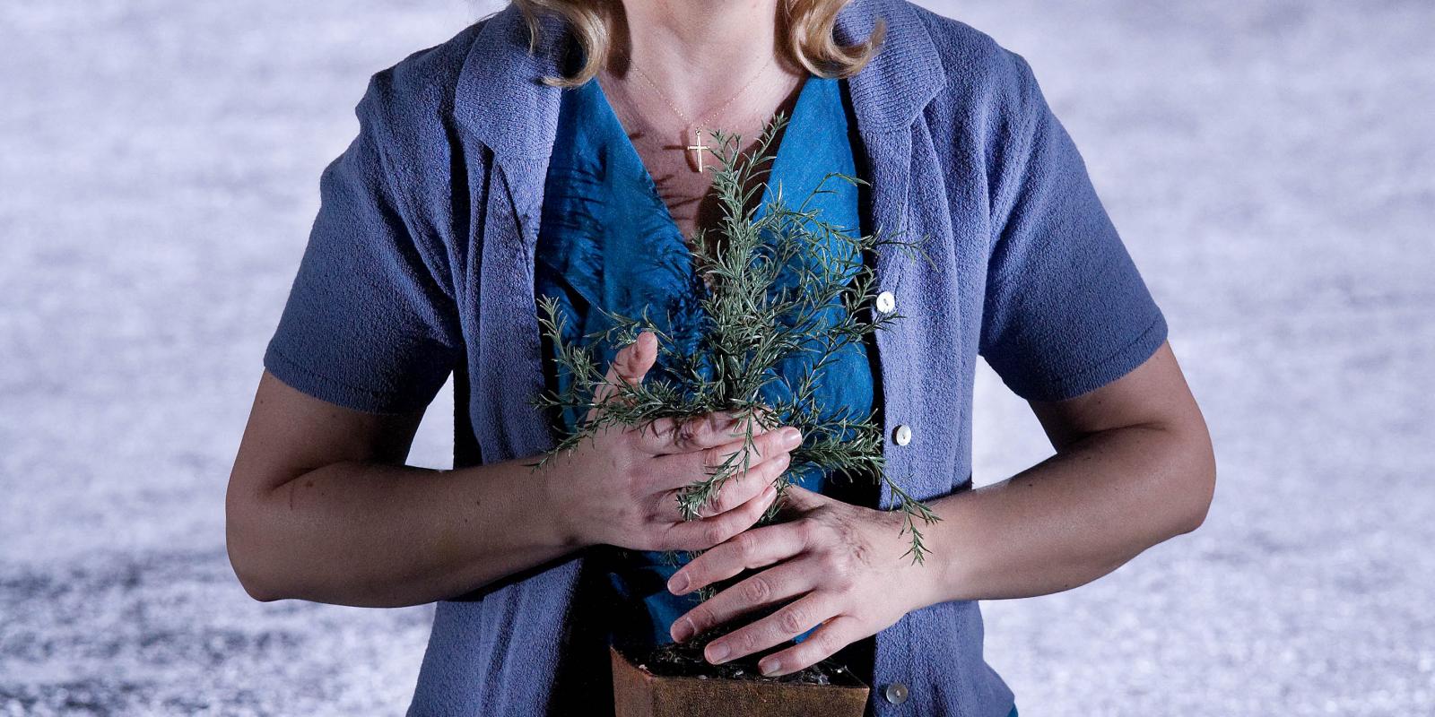 Woman in blue outfit holding a houseplant in ENO's Jenufa