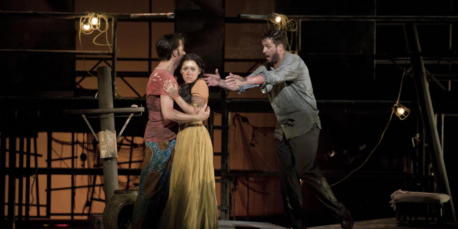 Man in a red shirt holds a woman in a yellow dress while another man rushes toward them in ENO's The Pearl Fishers