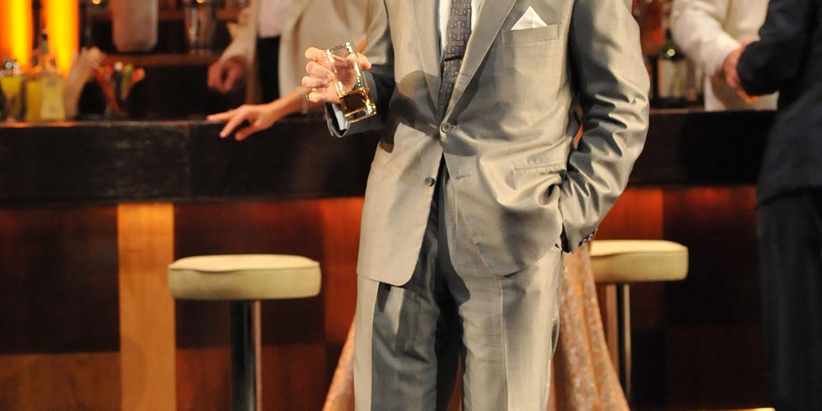 zoomed in photo of man in grey suit drinking whiskey from a tall glass in ENO's Rigoletto