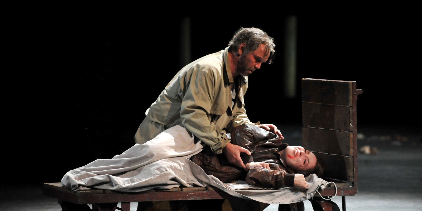 woman laying on wooden cart with man beside her on stage in rigoletto