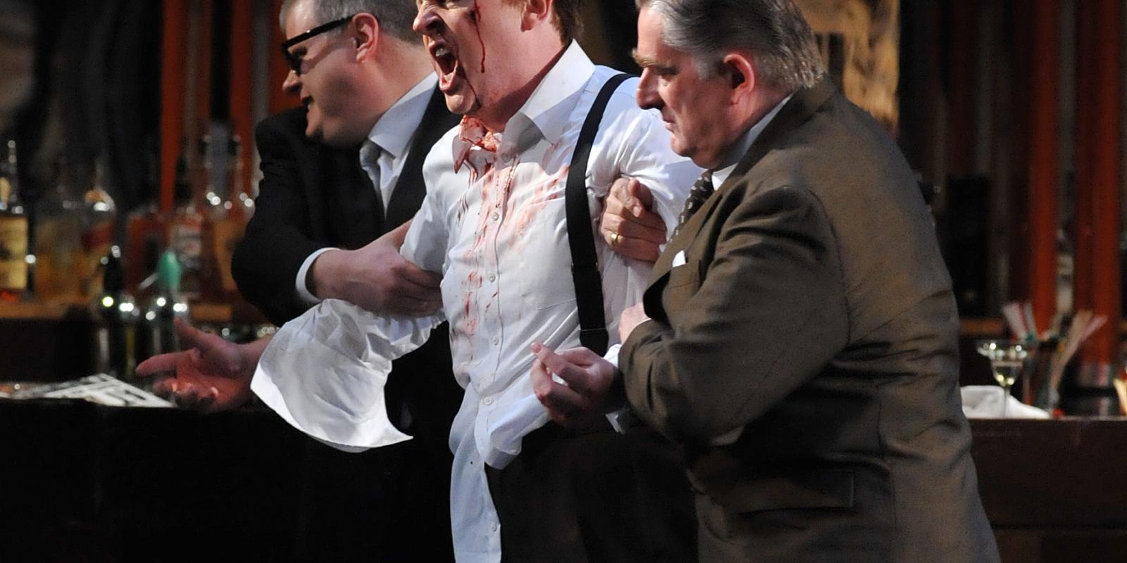 scene from ENO's Rigoletto of man covered in blood being held up by two other men