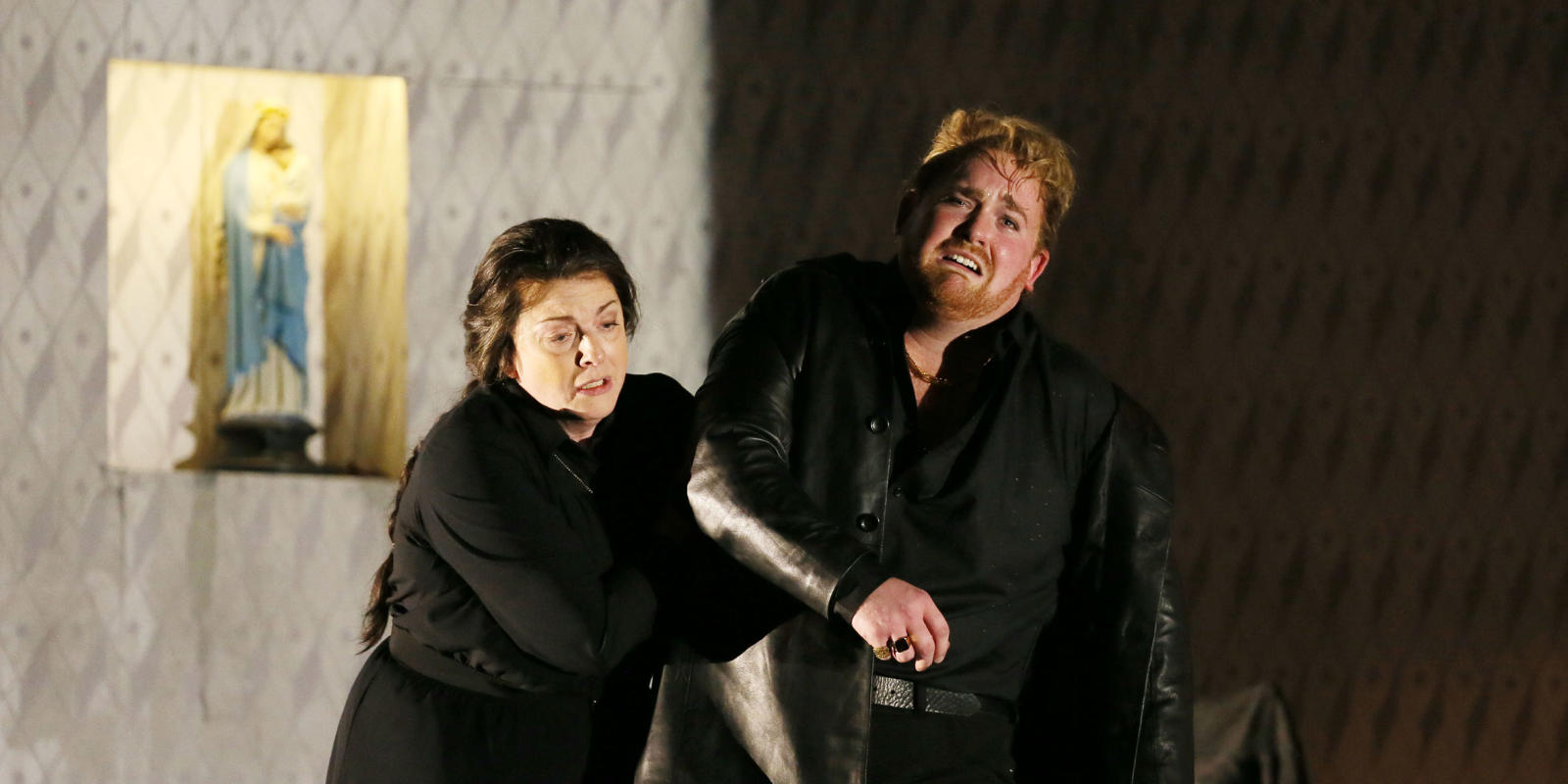 Visibly distraught man with ginger hair in a black coat being ushered by a woman in ENO's Jenufa