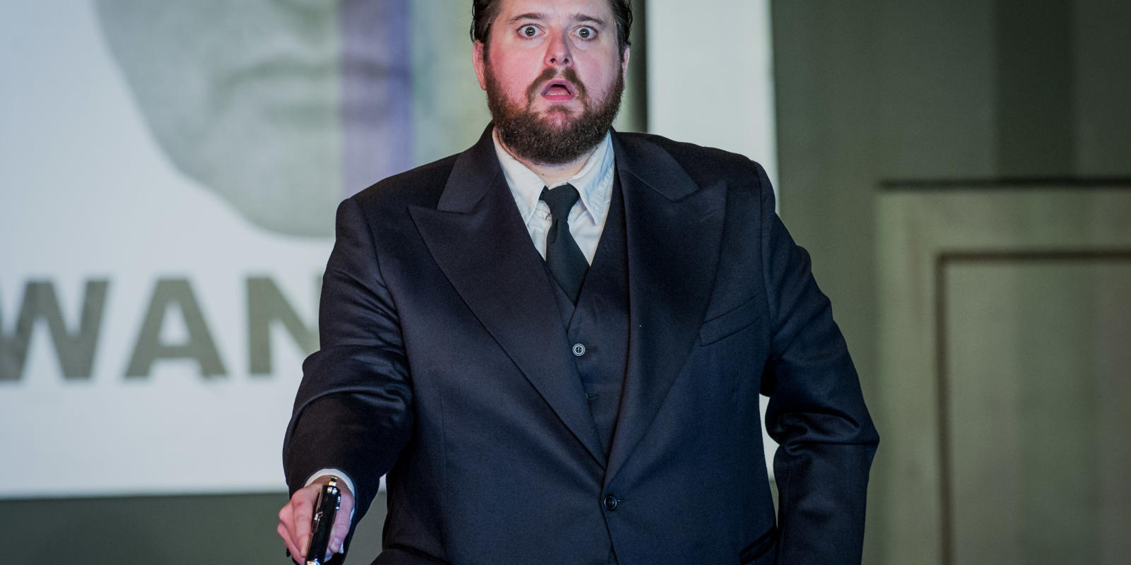 Bearded man wearing a dark suit whilst holding a gun in ENO's Don Giovanni