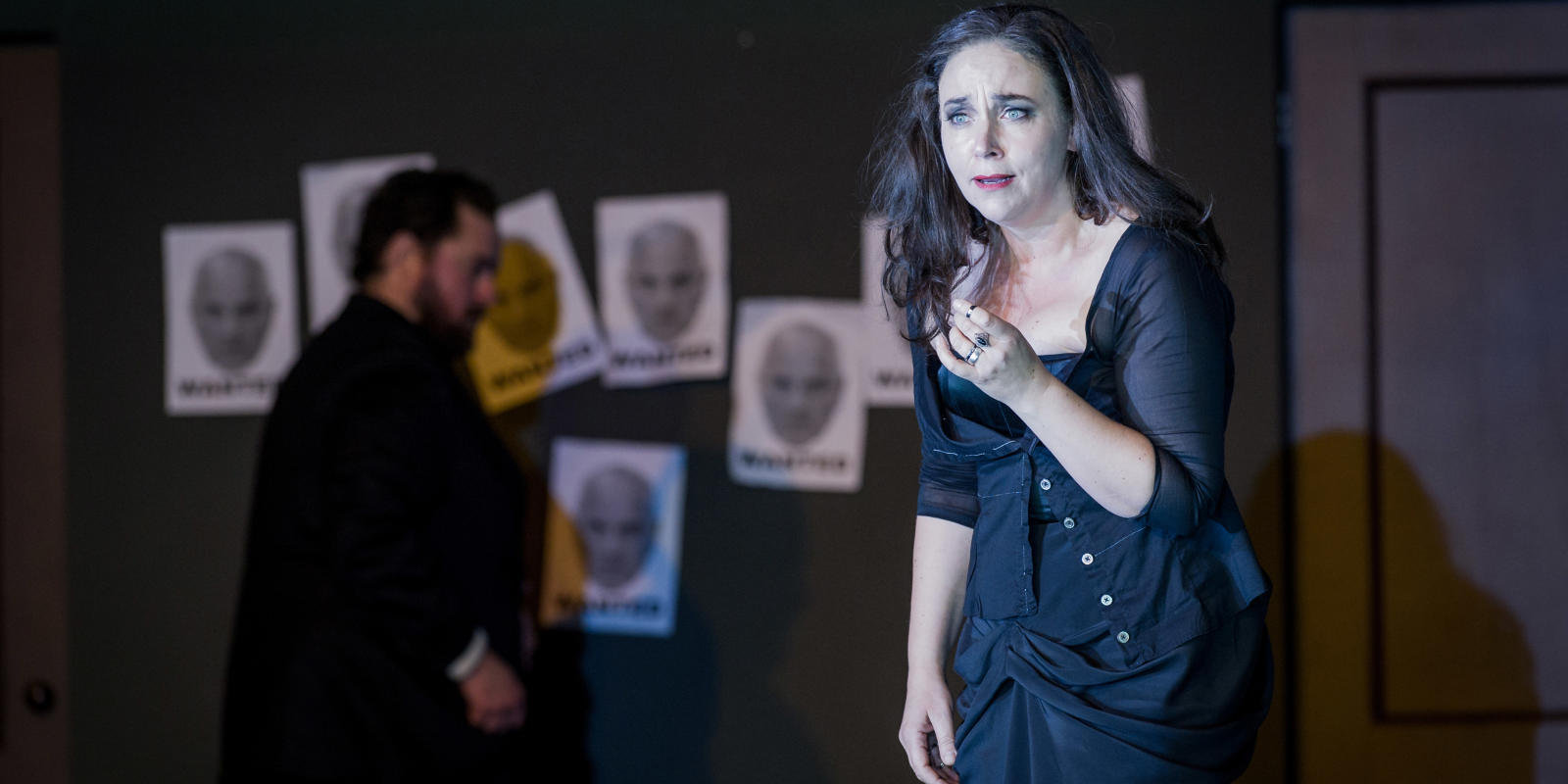 woman looking sad with man and posters with faces on them in the background in Don Giovanni