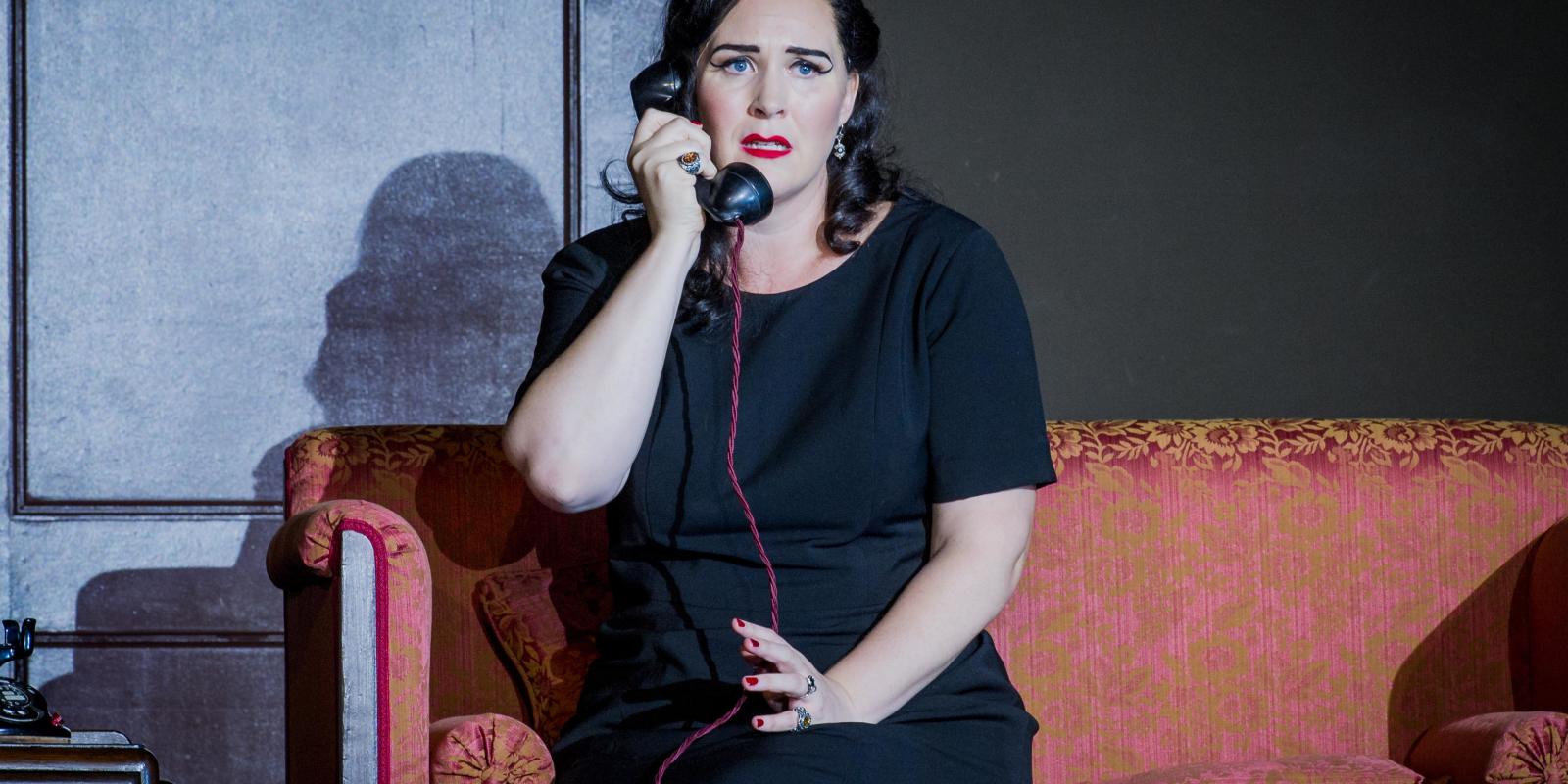 Woman sitting on a red sofa while speaking on the phone in ENO's Don Giovanni