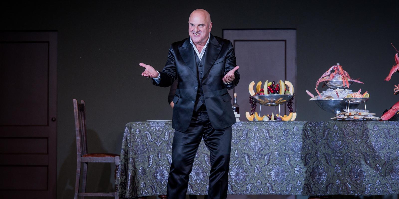 Man with no hair standing in front of a table of food, facing the audience with his hands open