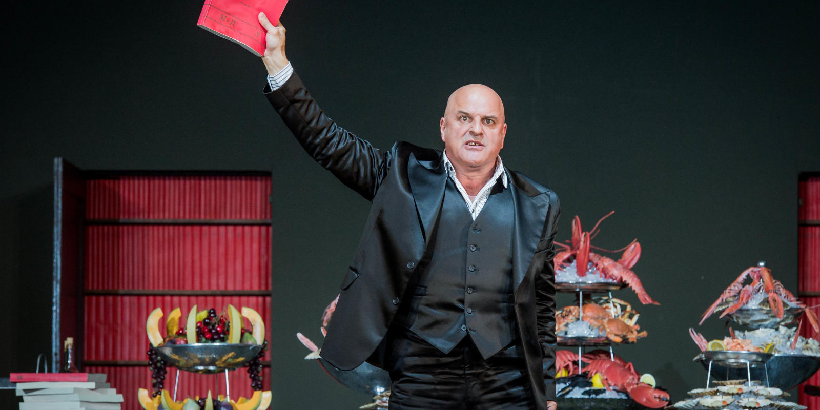 Bald man standing in front of a table of food while holding up a red book in ENO's Don Giovanni