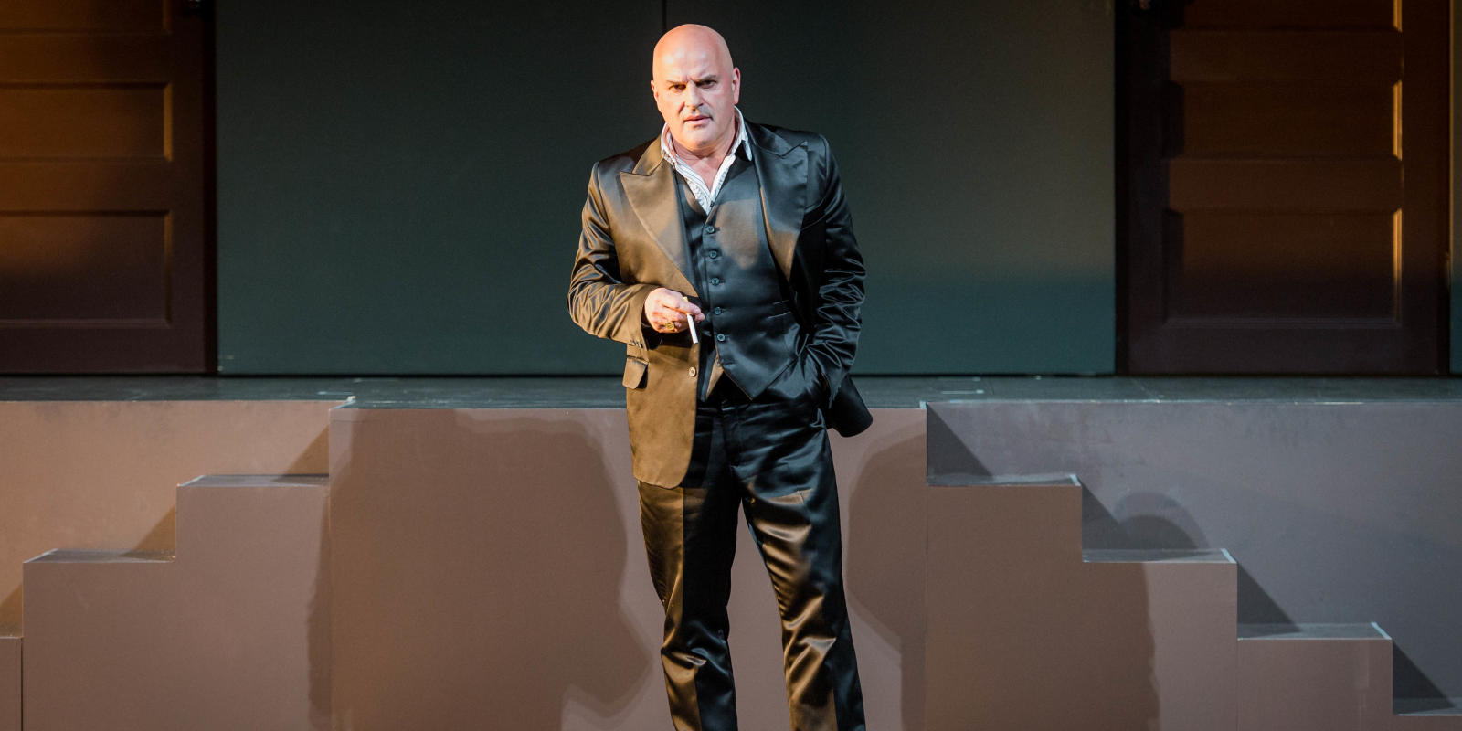 Man with no hair wearing an all black suit speaking toward the audience in ENO's Don Giovanni