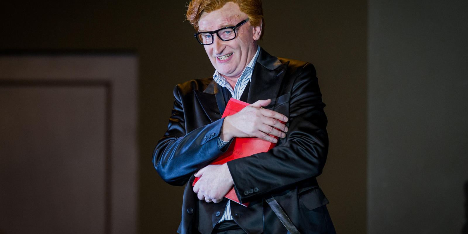 Ginger man with glasses and a black coat holding a red book in ENO's Don Giovanni