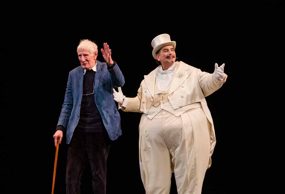 a man in a white suit and top hat, with an elderly gentlemen holding a walking stick. Both waving to the stage