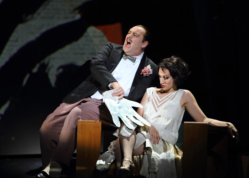 Man sitting next to a woman while singing and holding a large white glove in ENO's Lulu