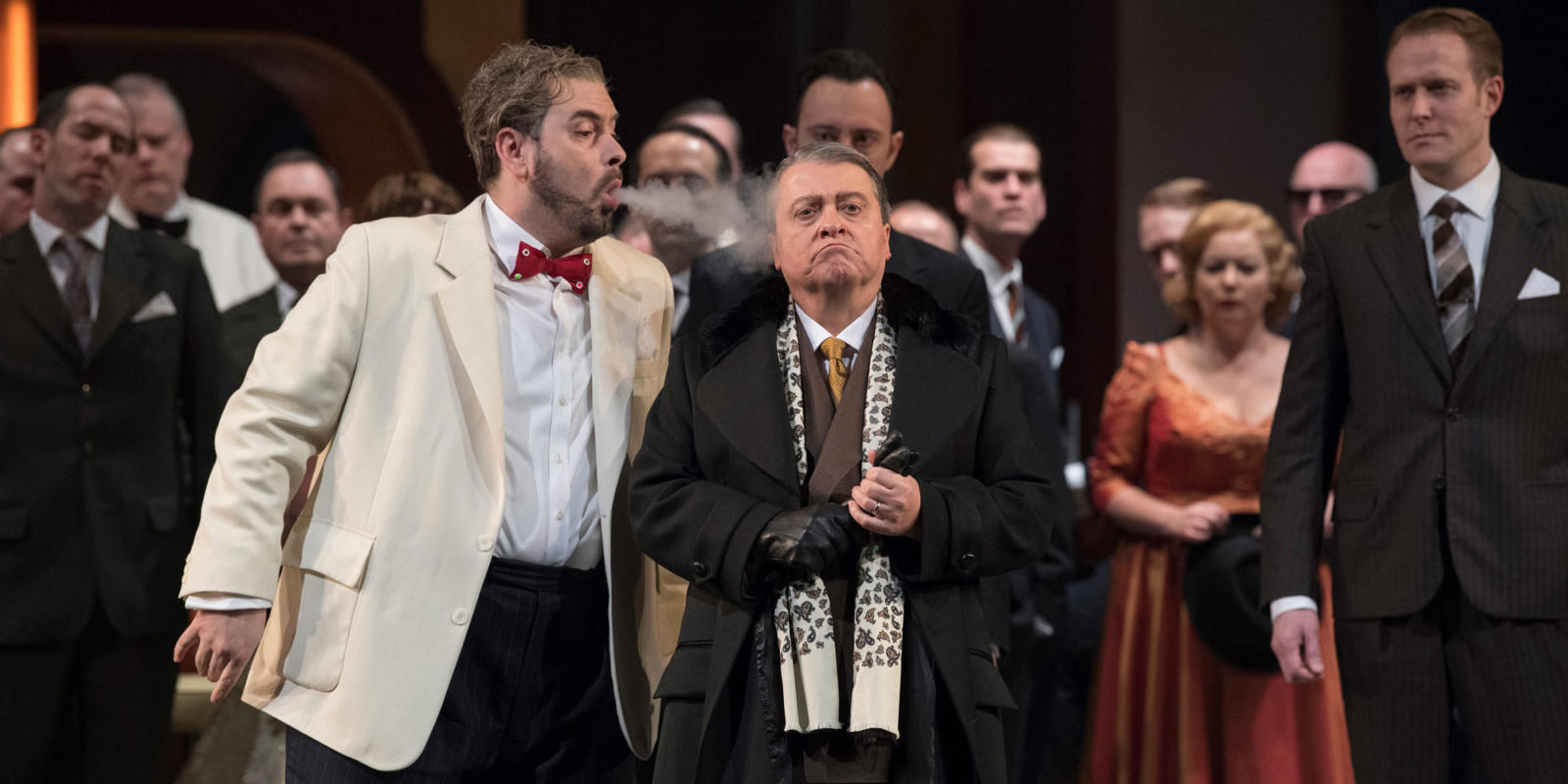 man in white suit blows smoke in the face of man in black suit on stage of Rigoletto