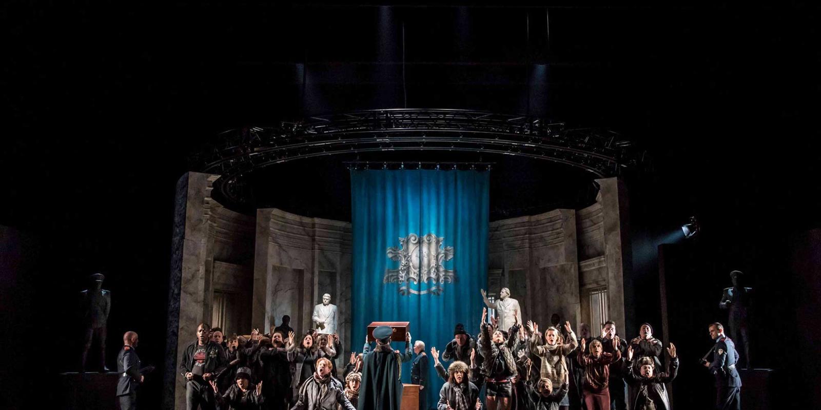 Group of people singing to the audience while a man holds a box in the air in ENO's The Winter's Tale