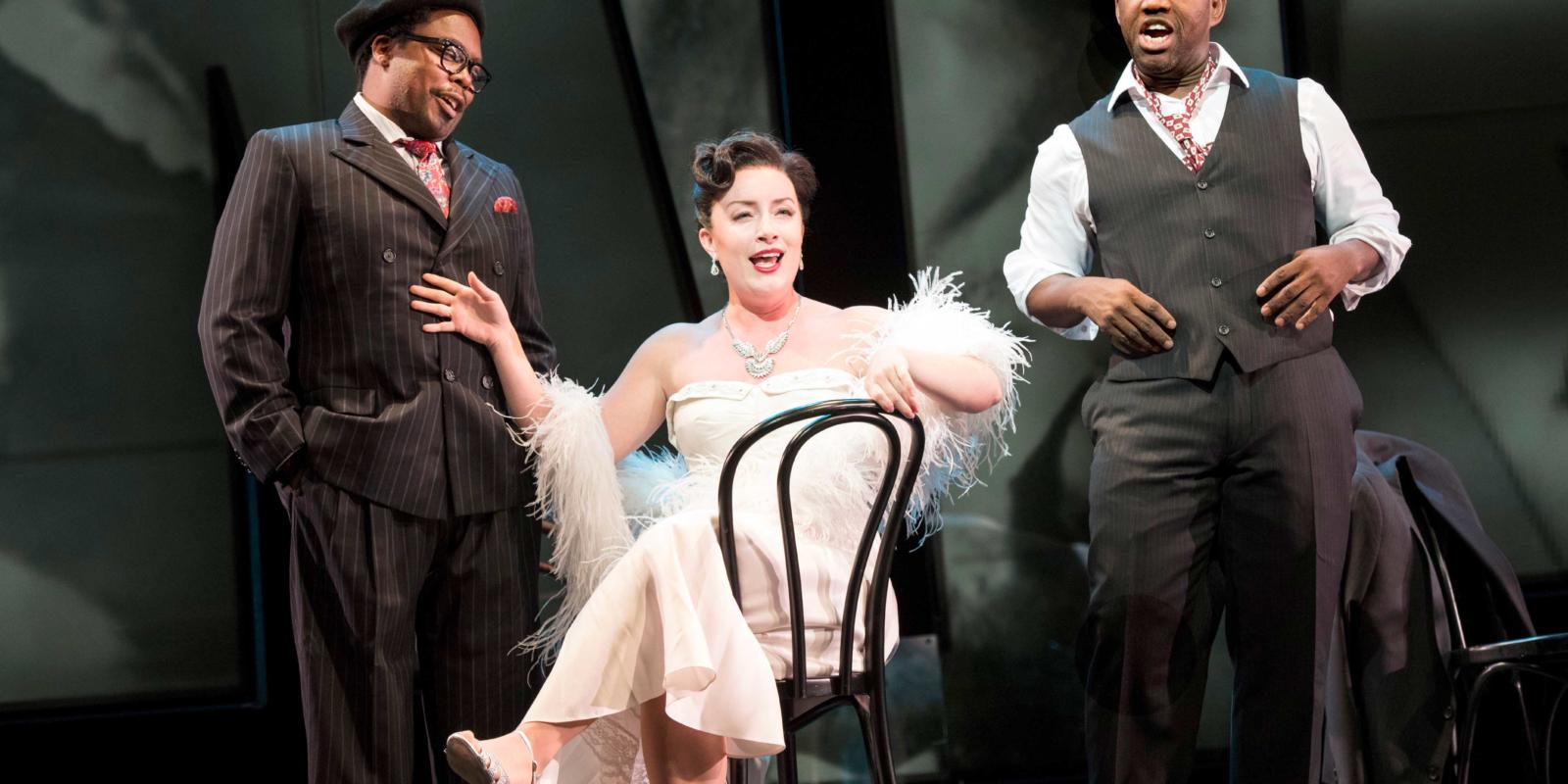 a women in a white dress sat on a chair singing to two men dressed in suits