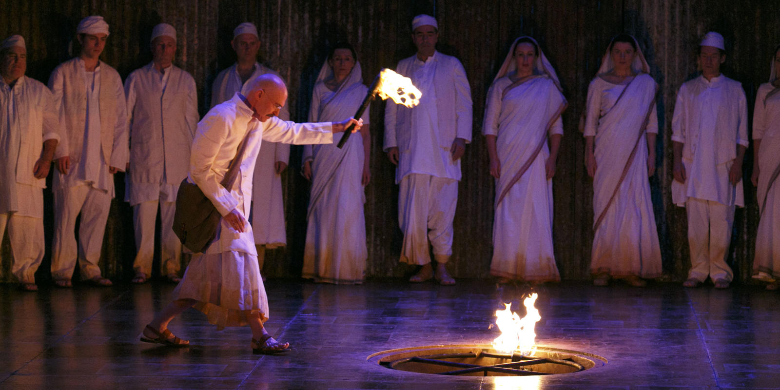 Man with a torch looking into a pit, whilst surrounded by a group in ENO's Satyagraha