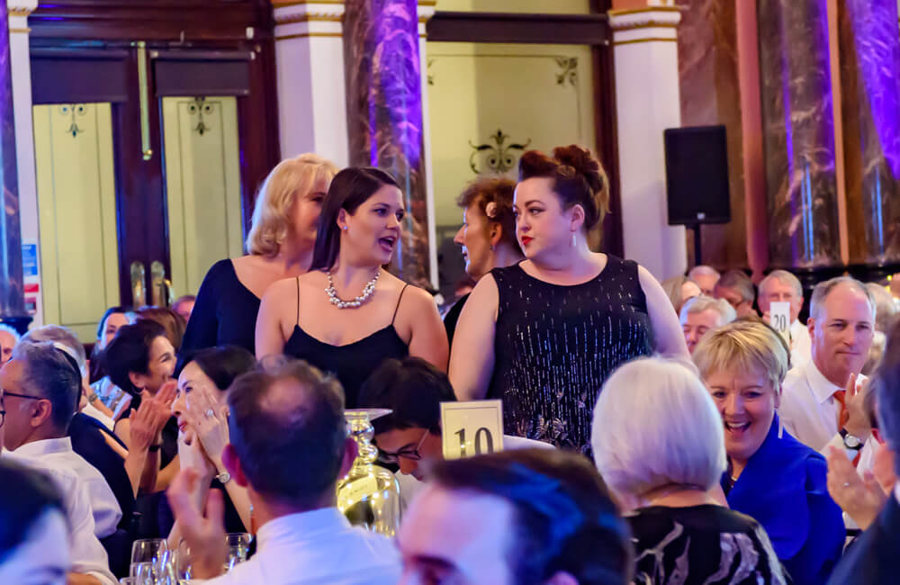 two women in black dresses standing amongst people sitting at tables at ENO's gala dinner