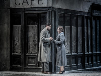 Mimi and Rodolfo in La Bohème clutching each other's hands in front of a shop