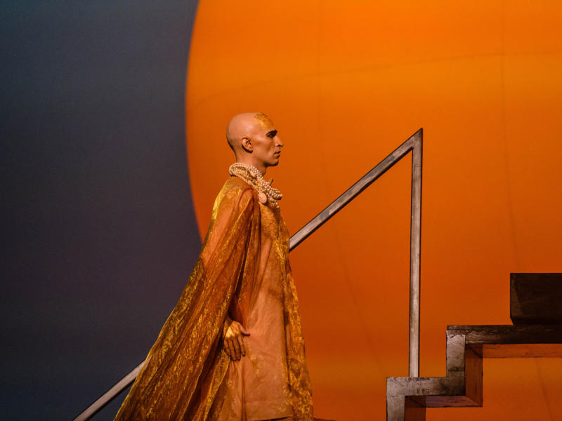 Anthony Roth Costanzo in the title role of Akhaten wearing a velvet orange cape in front of the bright orange sun