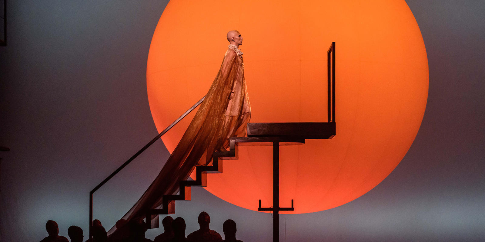 Anthony Roth Costanzo in the title role of Akhaten wearing a velvet orange cape in front of the bright orange sun