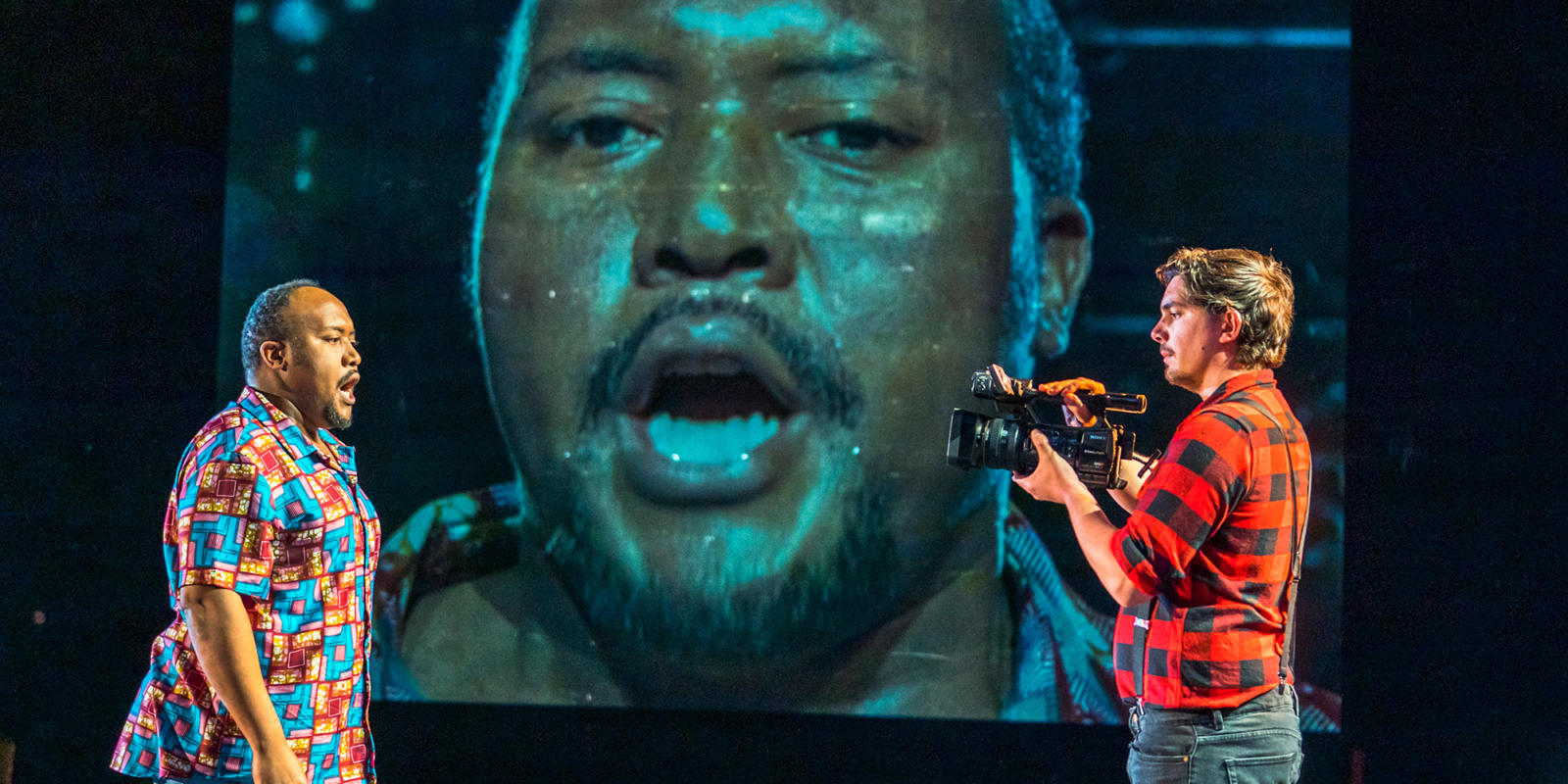 a man singing at a camera man on stage, a projection of the screen is behind them