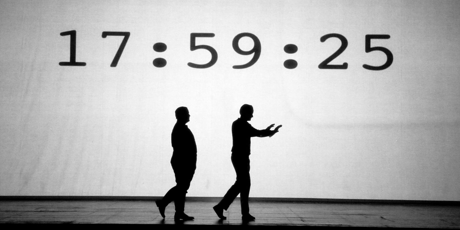 Orphée and Heurtebise walk silhouetted against a white background, with a bold timecode projected onto it.