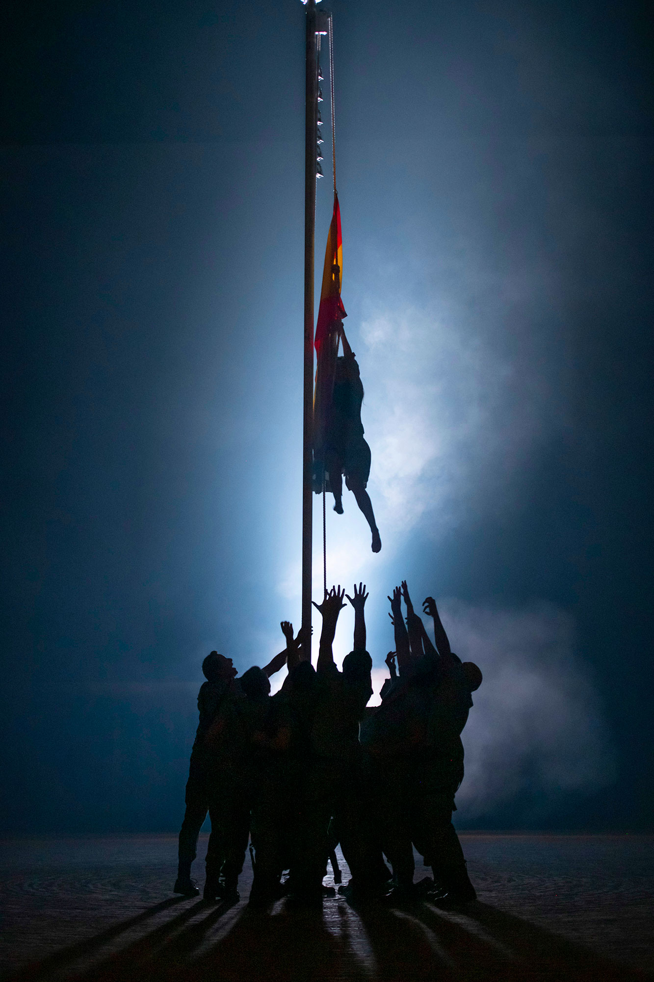 Silhouetted group stand around a flagpole, reaching up for the Spanish flag