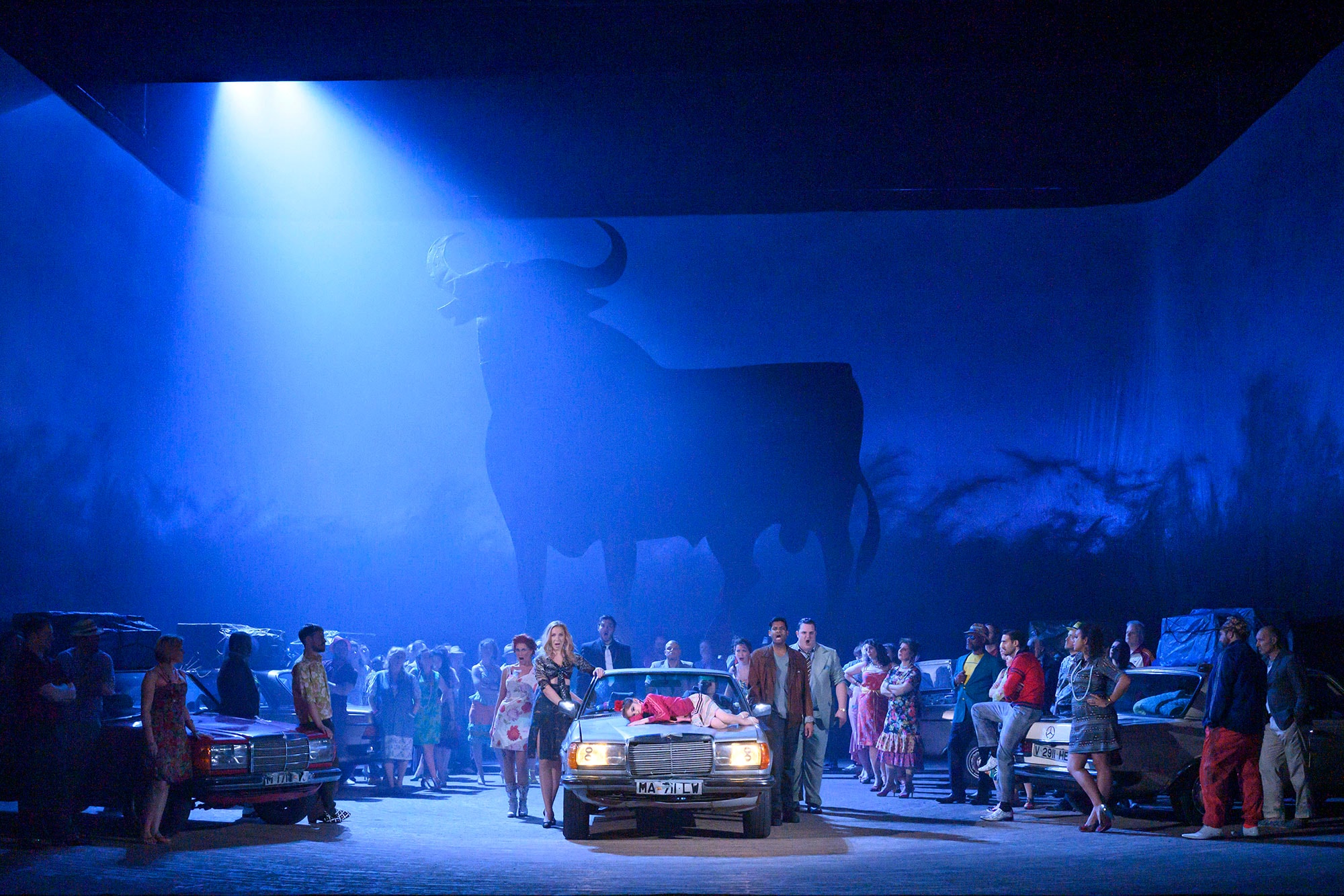 ENO Carmen 2020, ensemble shot waround cars, with silhouette of bull in background