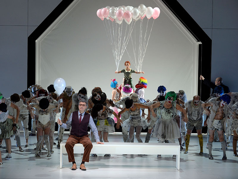 ENO chorus on stage with a person at the back attached to balloons