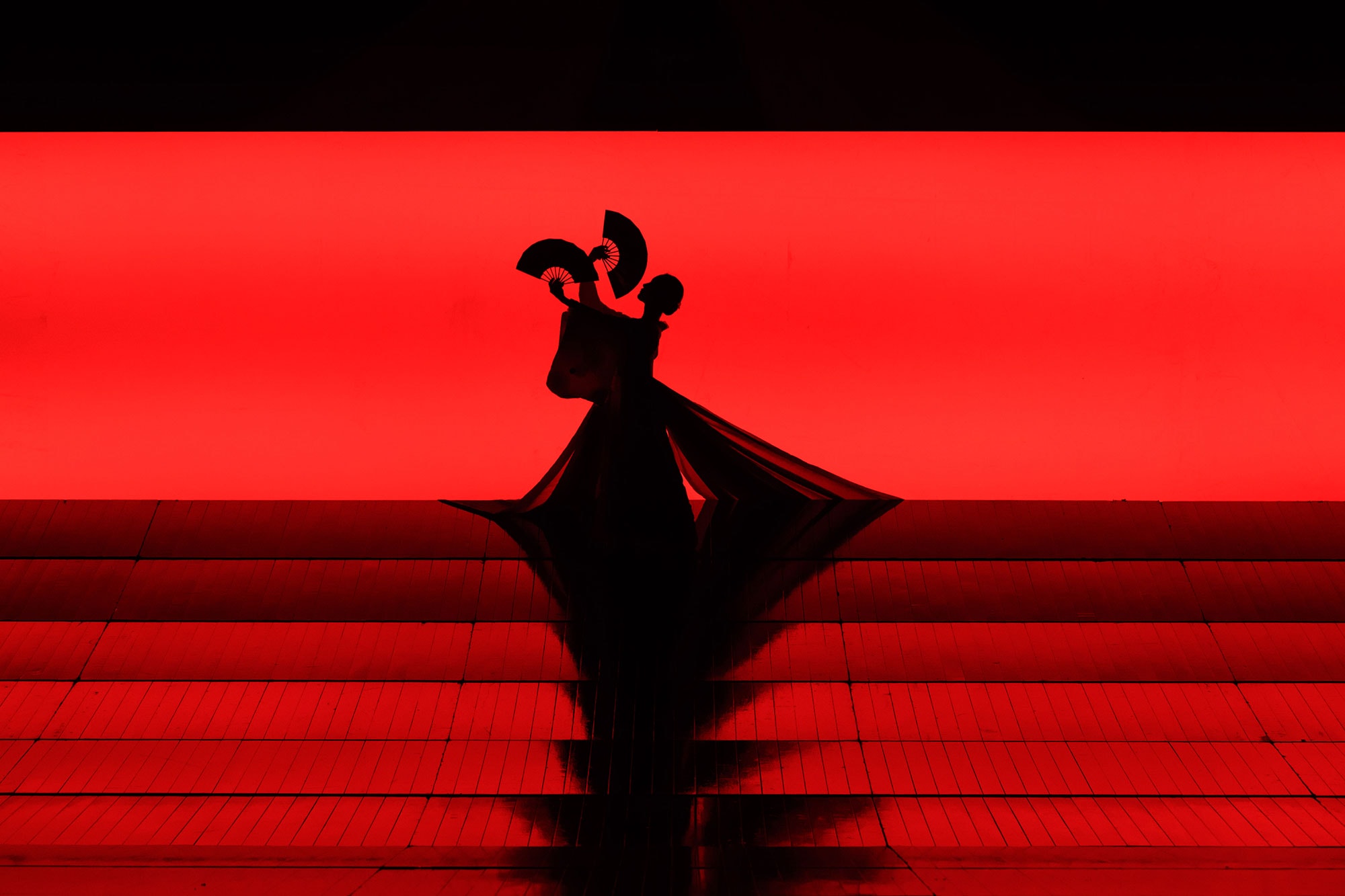 Fan dancer stands silhouetted against a red background at the top of the stage