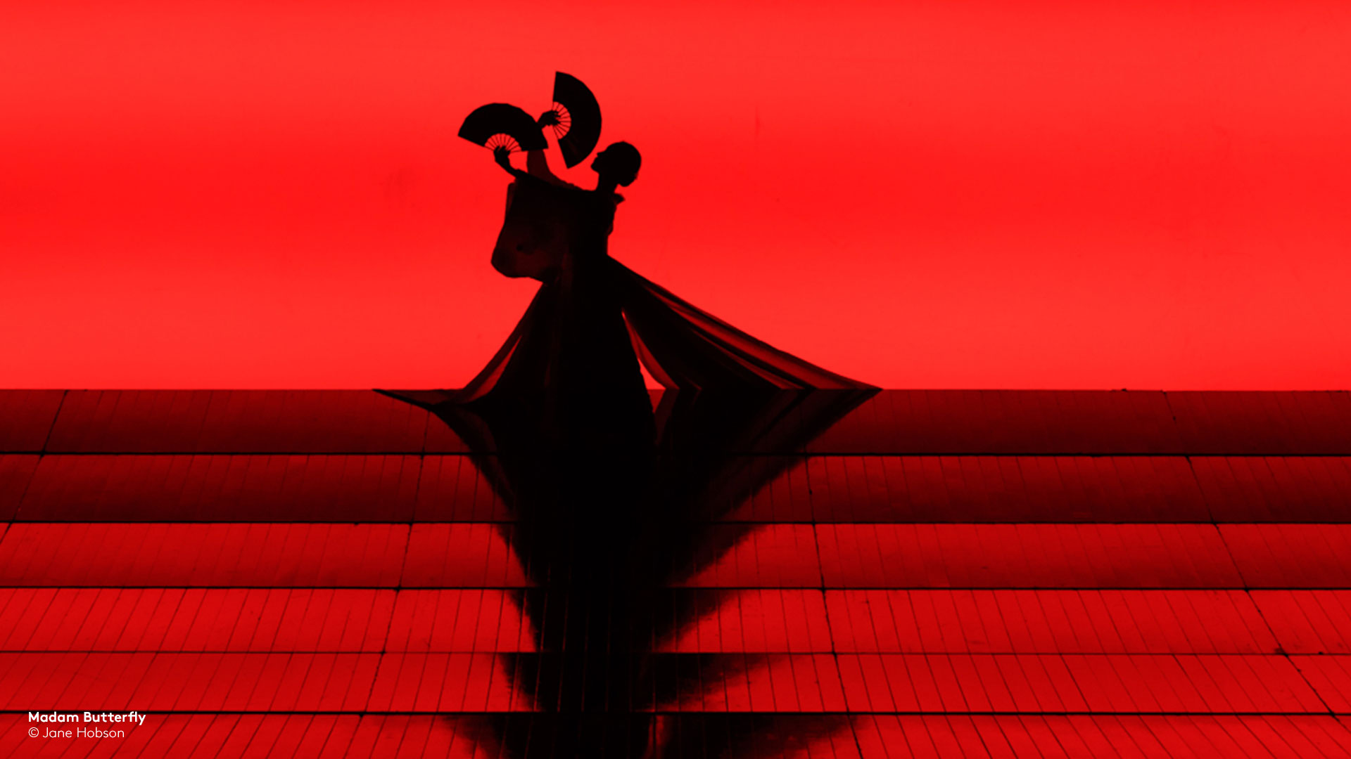 silhouette from ENO's Madam Butterfly of woman holding two fans performing on red stage