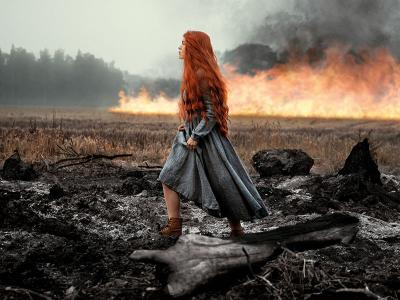 woman with long ginger hair walking through a field with fire in the background