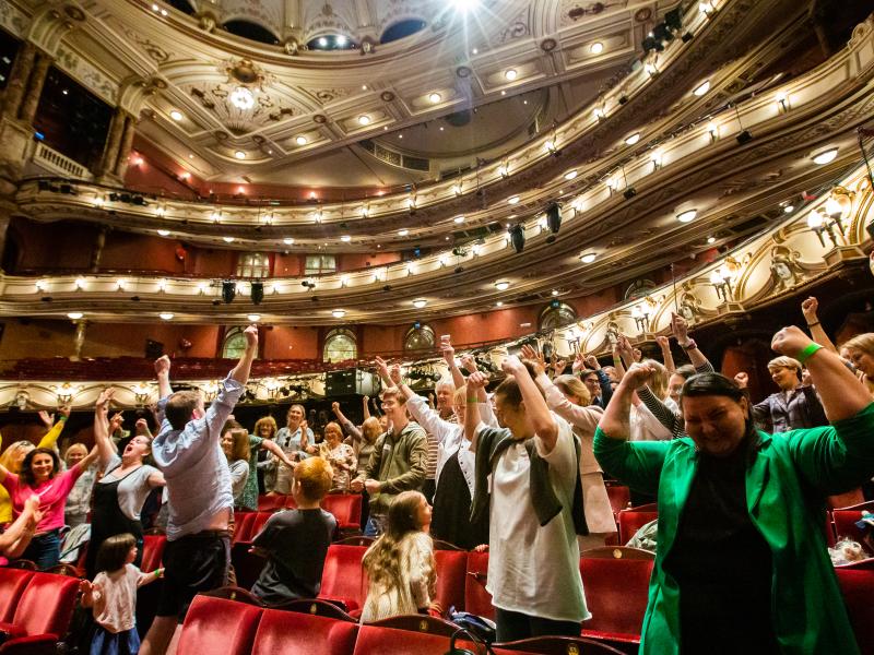 Large number of people in workshop with arms raised in London Coliseum auditorium