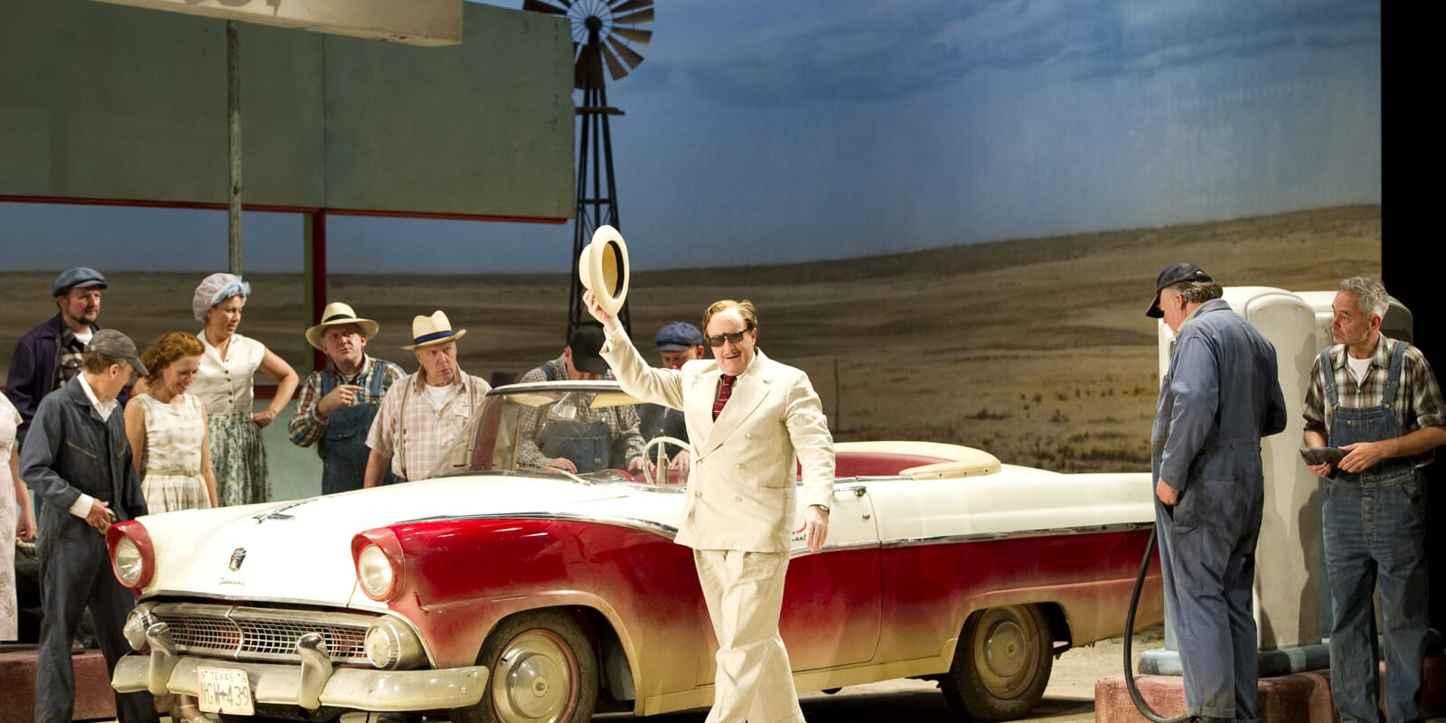 A man in a cream suit waves his hat in the air while standing by a red car