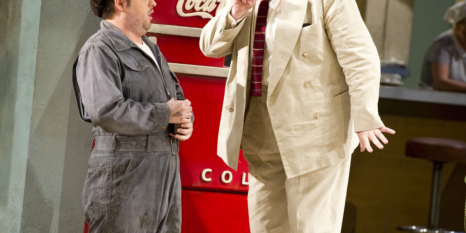 A man in a cream suit interacts with a man in a grey boiler suit and jacket by a vending machine