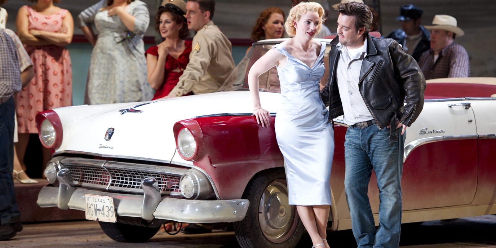 A man in a black leather jacket and a woman in a blue dress stand next to a cream and red car