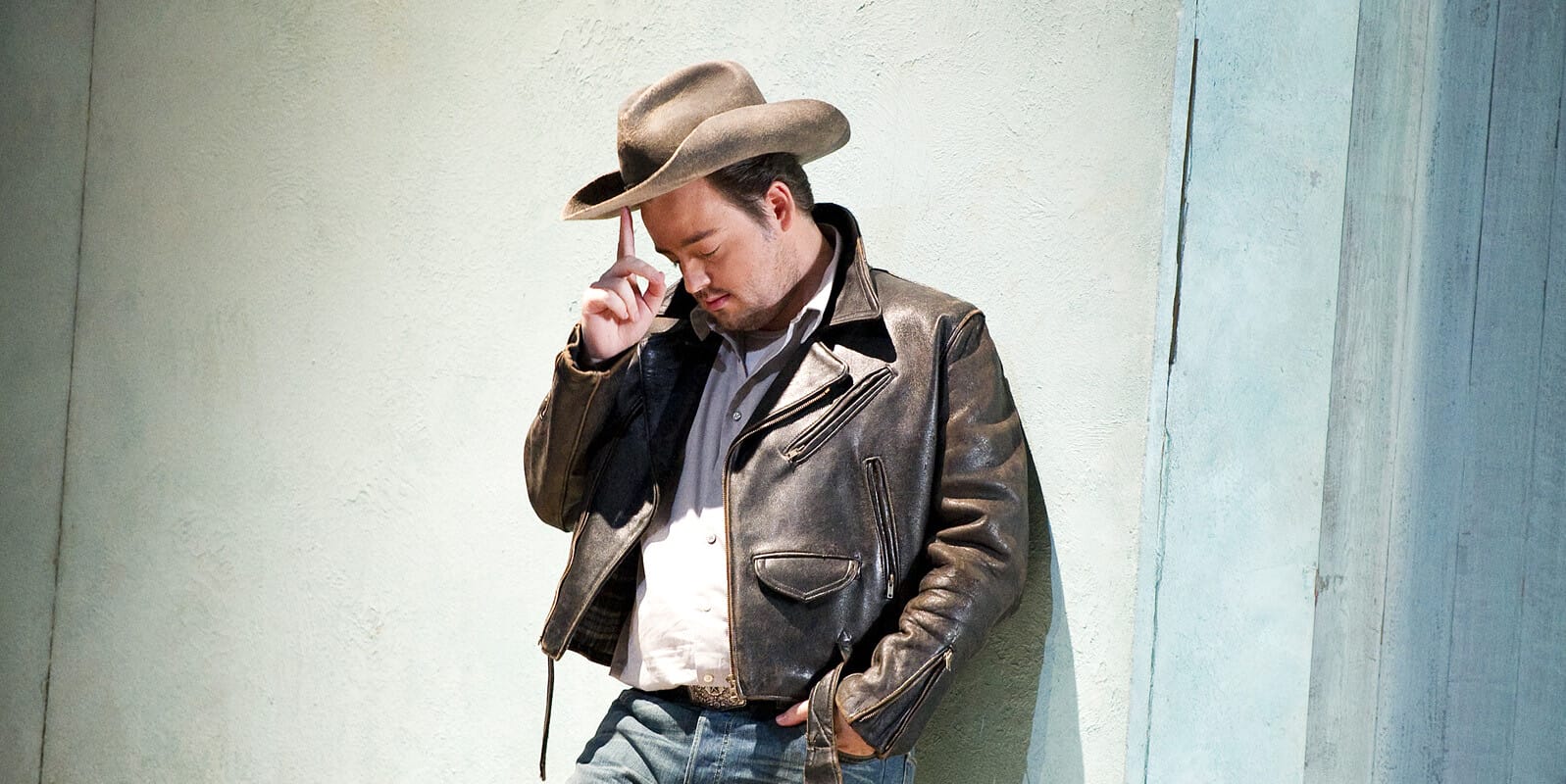 Man in cowboy hat and leather jacket leans against wall