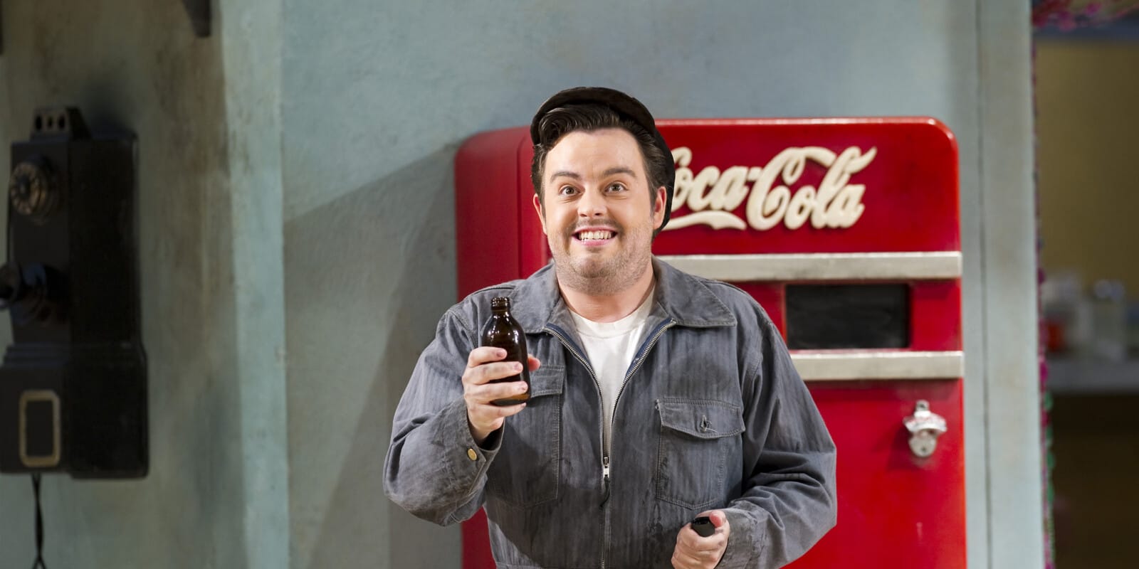 A man in a grey boiler suit stands next to a vending machine smiling.