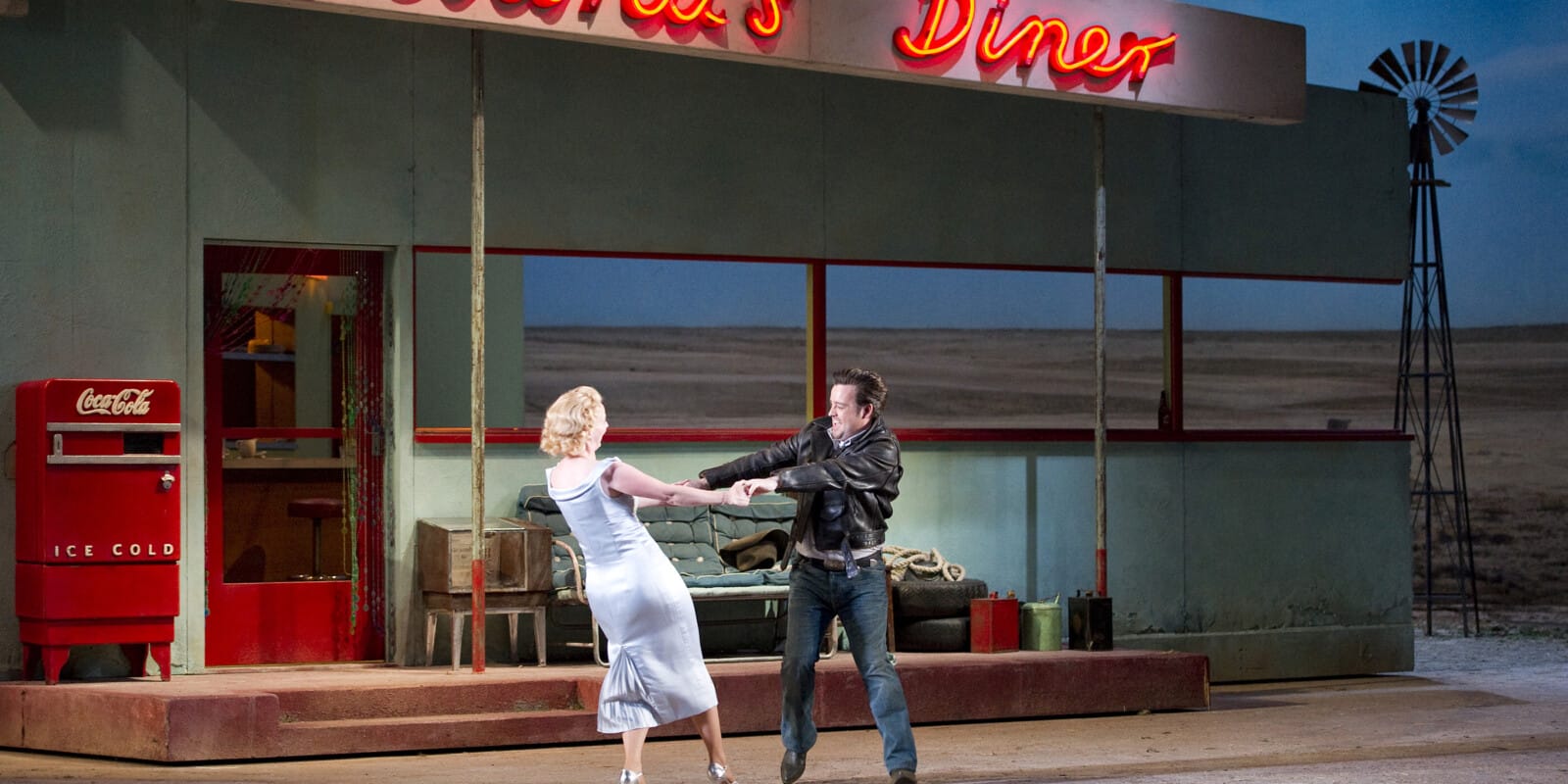 Woman in blue dress and man in black leather jacket dance underneath Adina's Diner sign